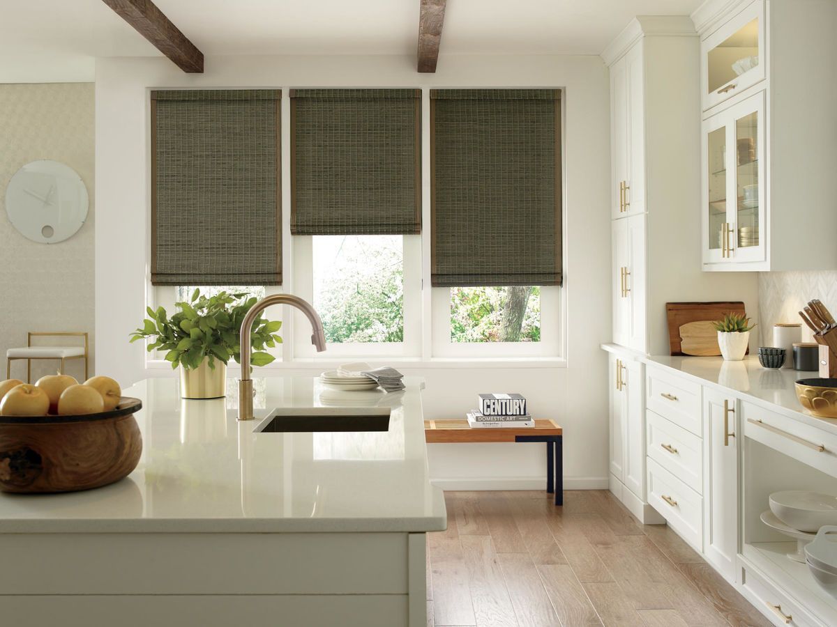 Kitchen with white cabinets, island, and woven shades. Gold faucet, wooden beams, and a plant on the island.