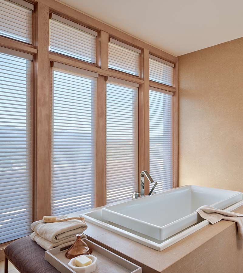 Bathroom with rectangular tub, large windows with white blinds, neutral wall, towels, and bath accessories.