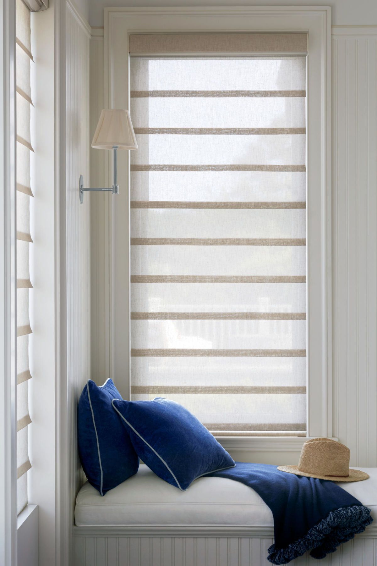 Window seat with blue pillows, straw hat, and banded neutral shade.