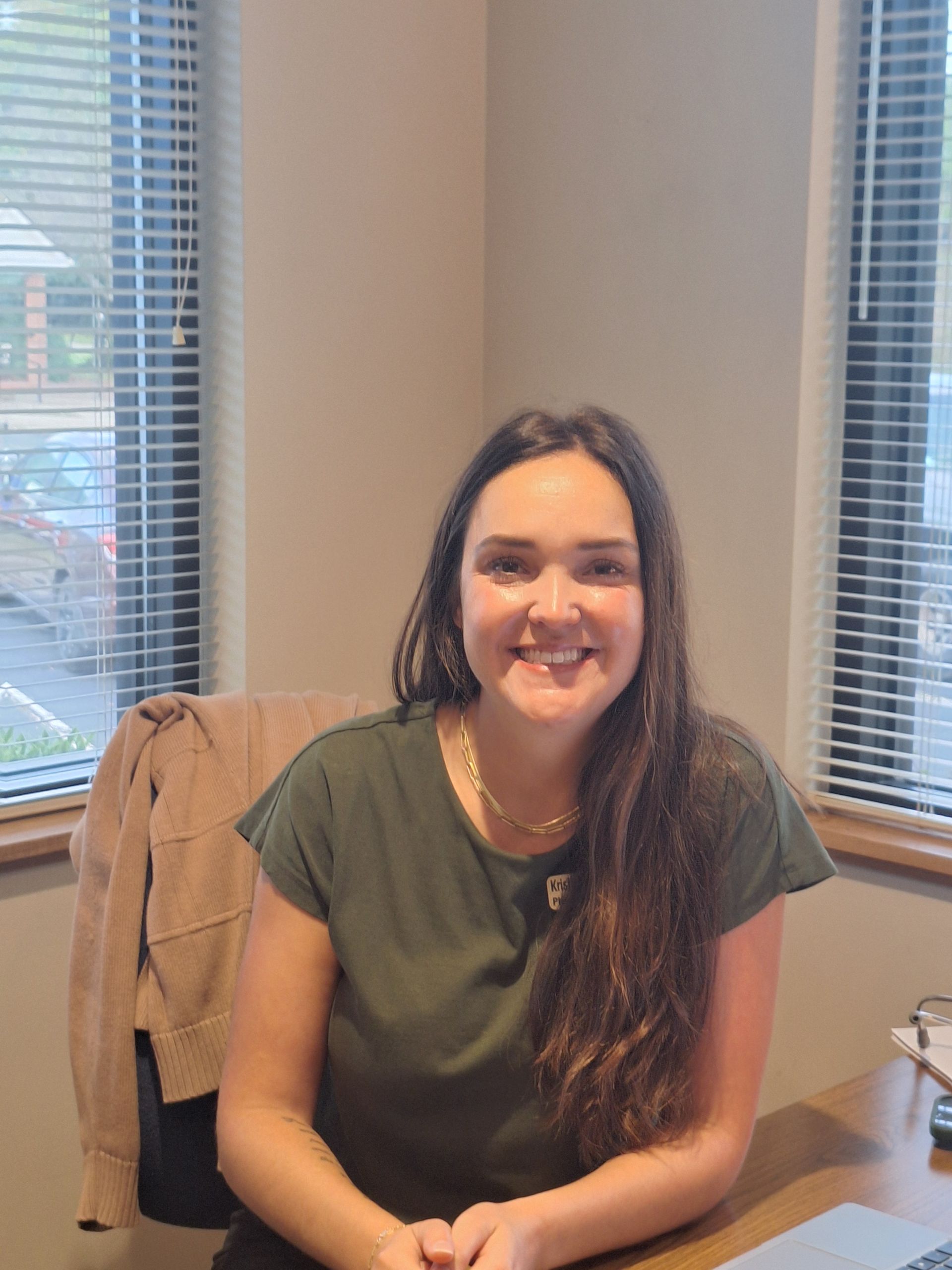 Woman with long brown hair smiling, seated at a desk, wearing green shirt. Window in background. Woman with long brown hair smiling, seated at a desk, wearing green shirt. Window in background.