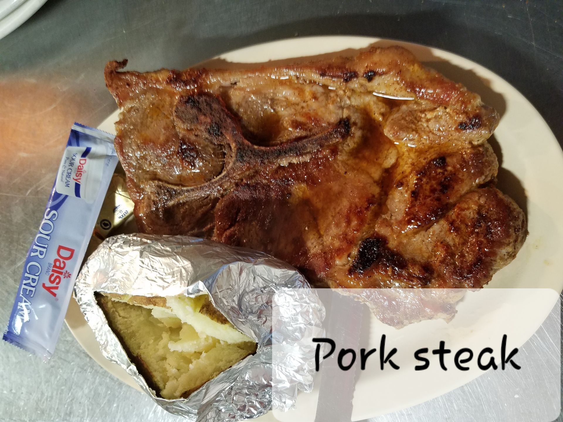 A cooked pork steak served on a plate with a baked potato wrapped in foil and a packet of Daisy sour cream.