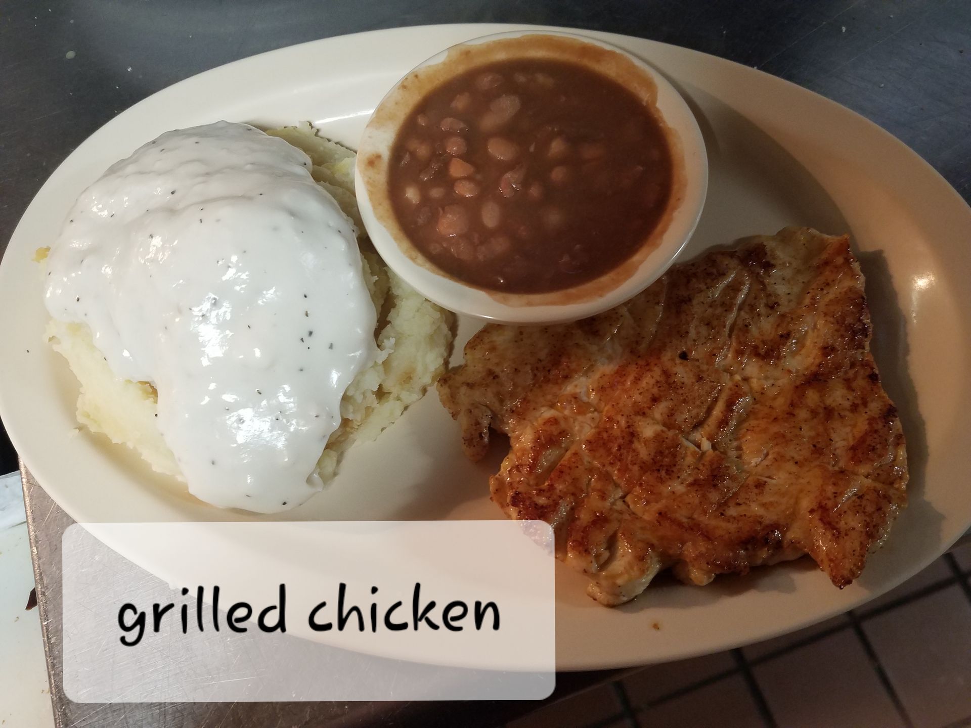 A meal on an oval plate featuring a grilled chicken breast, mashed potatoes topped with white gravy, and a side of beans.