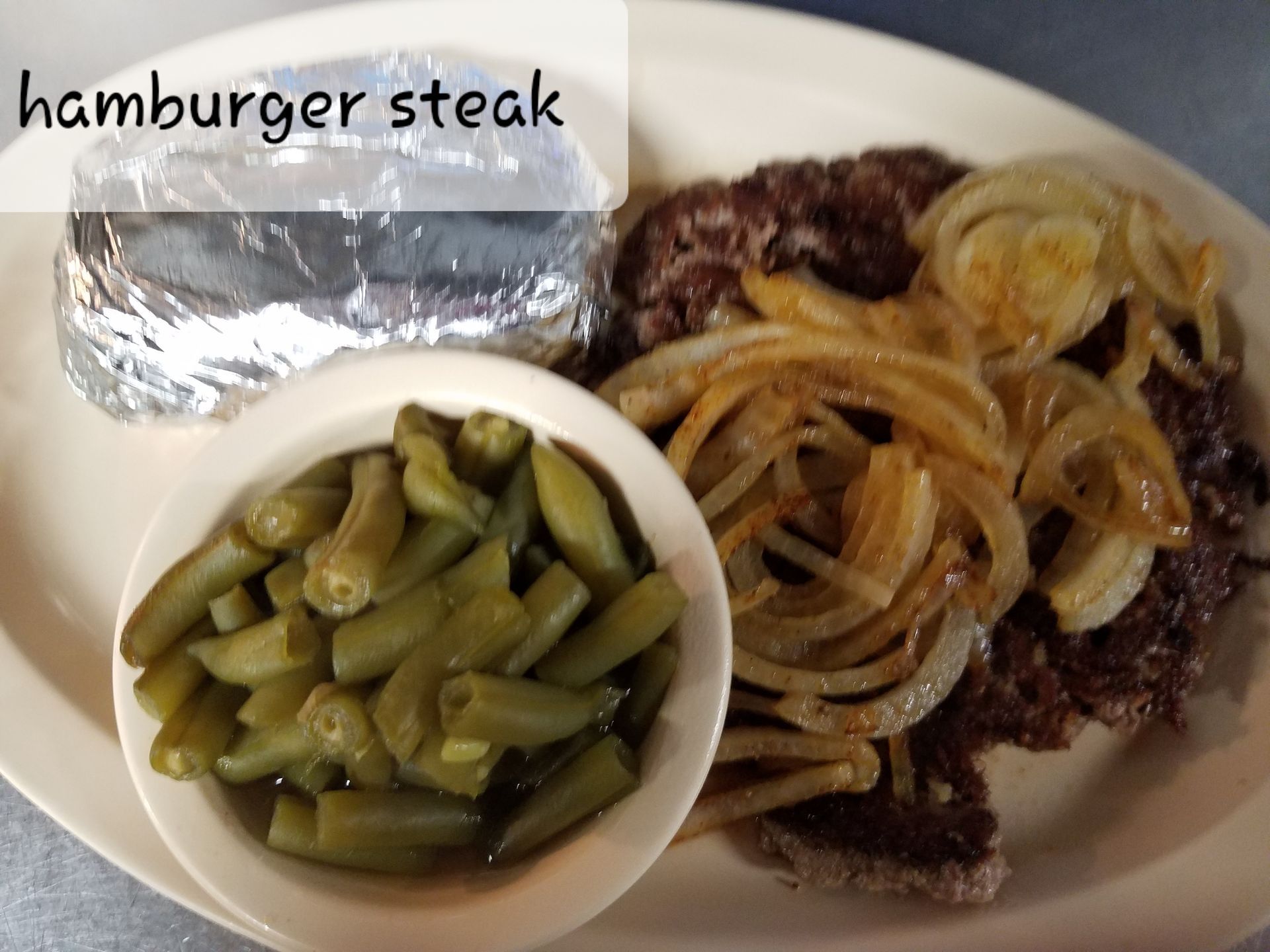A plate with a hamburger steak topped with onions, a bowl of green beans, and a foil-wrapped baked potato.