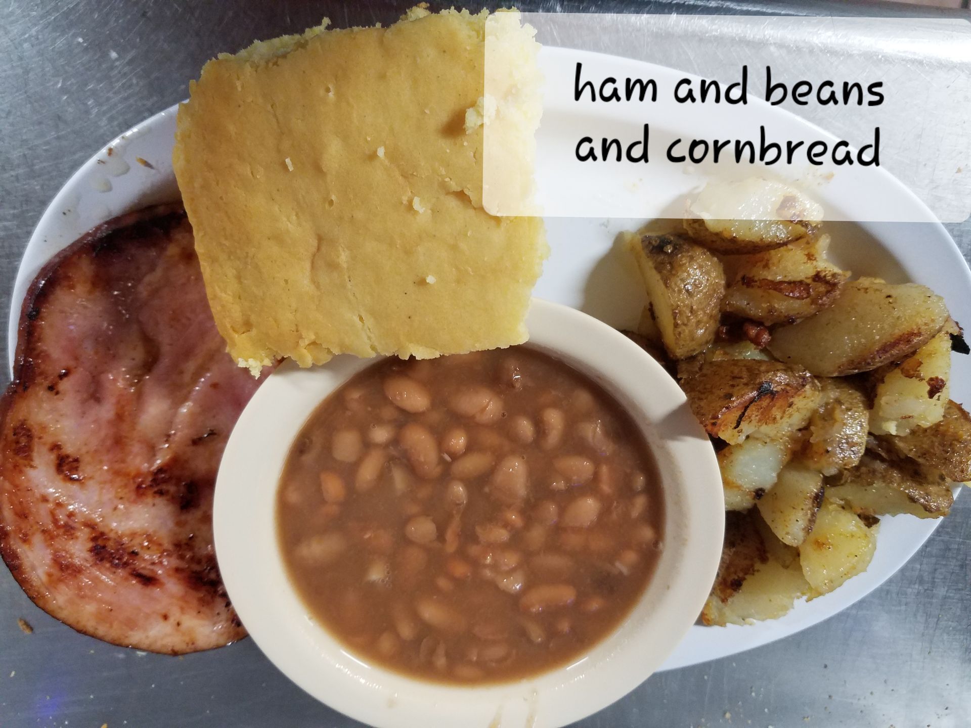 A plate containing a slice of ham, a bowl of brown beans, a square of yellow cornbread, and a side of roasted potatoes.
