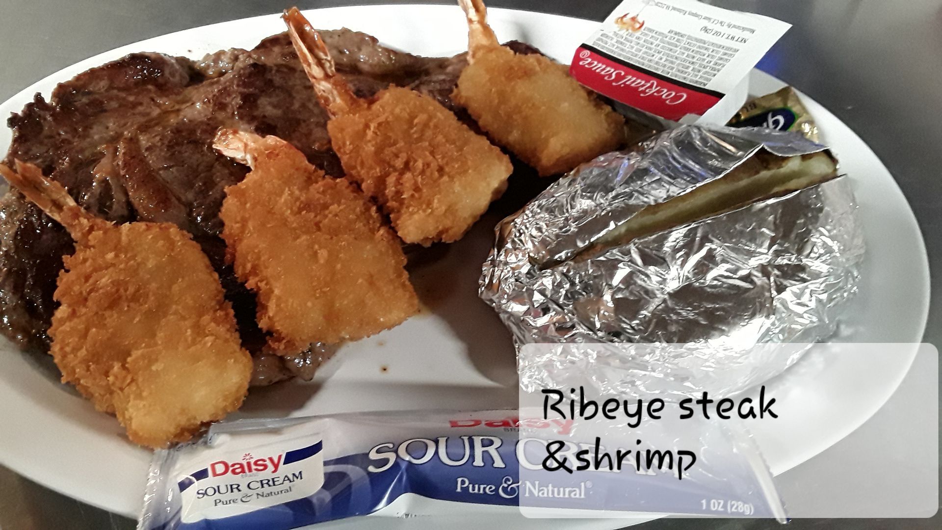 A ribeye steak topped with three fried shrimp, served with a baked potato in foil and packets of sour cream and butter.