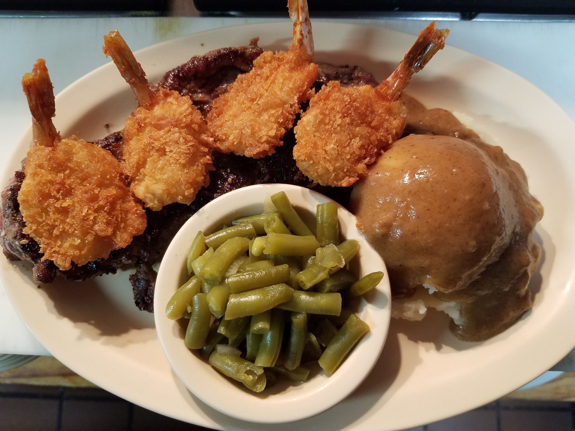 A plate with a steak topped with four breaded shrimp, a side of green beans, and mashed potatoes with gravy.