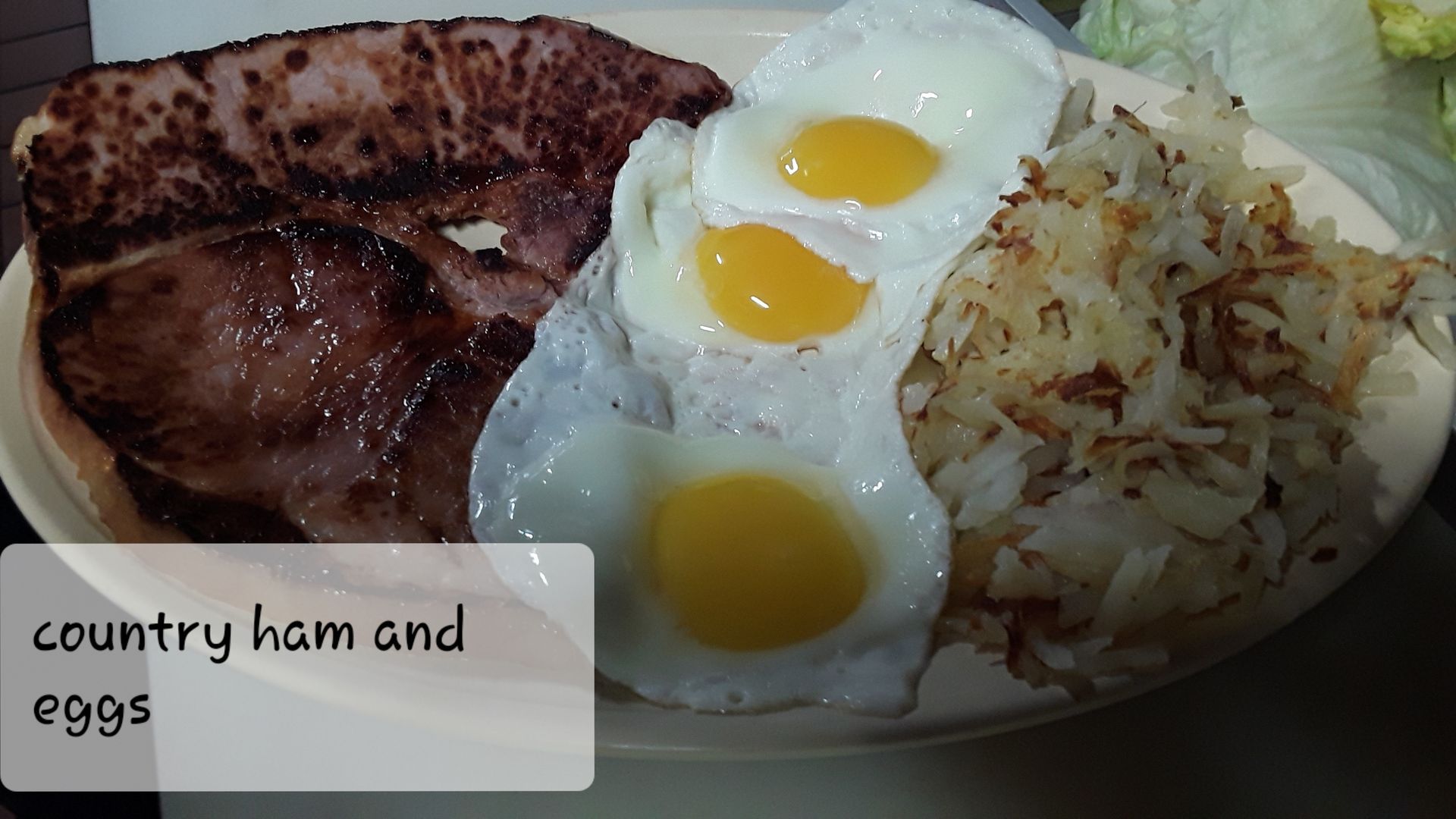 A breakfast plate with a large slice of grilled country ham, three sunny-side-up eggs, and a serving of hash browns.