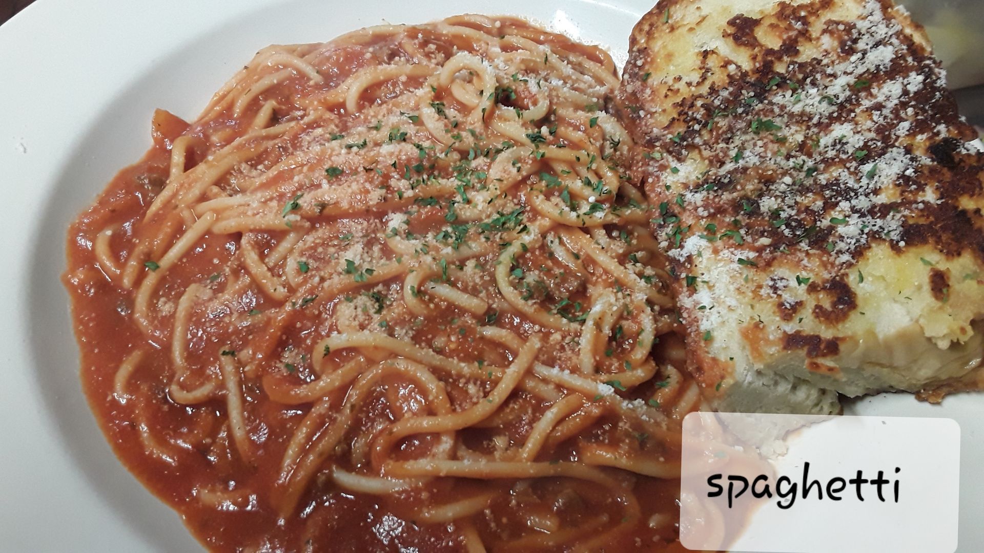 A plate of spaghetti with red sauce, topped with herbs and cheese, served with a piece of toasted bread.