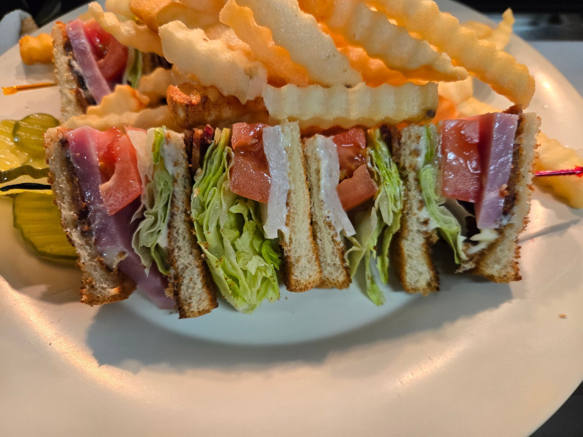 A club sandwich with ham, lettuce, and tomato, served with a side of crinkle-cut fries and pickles on a white plate.