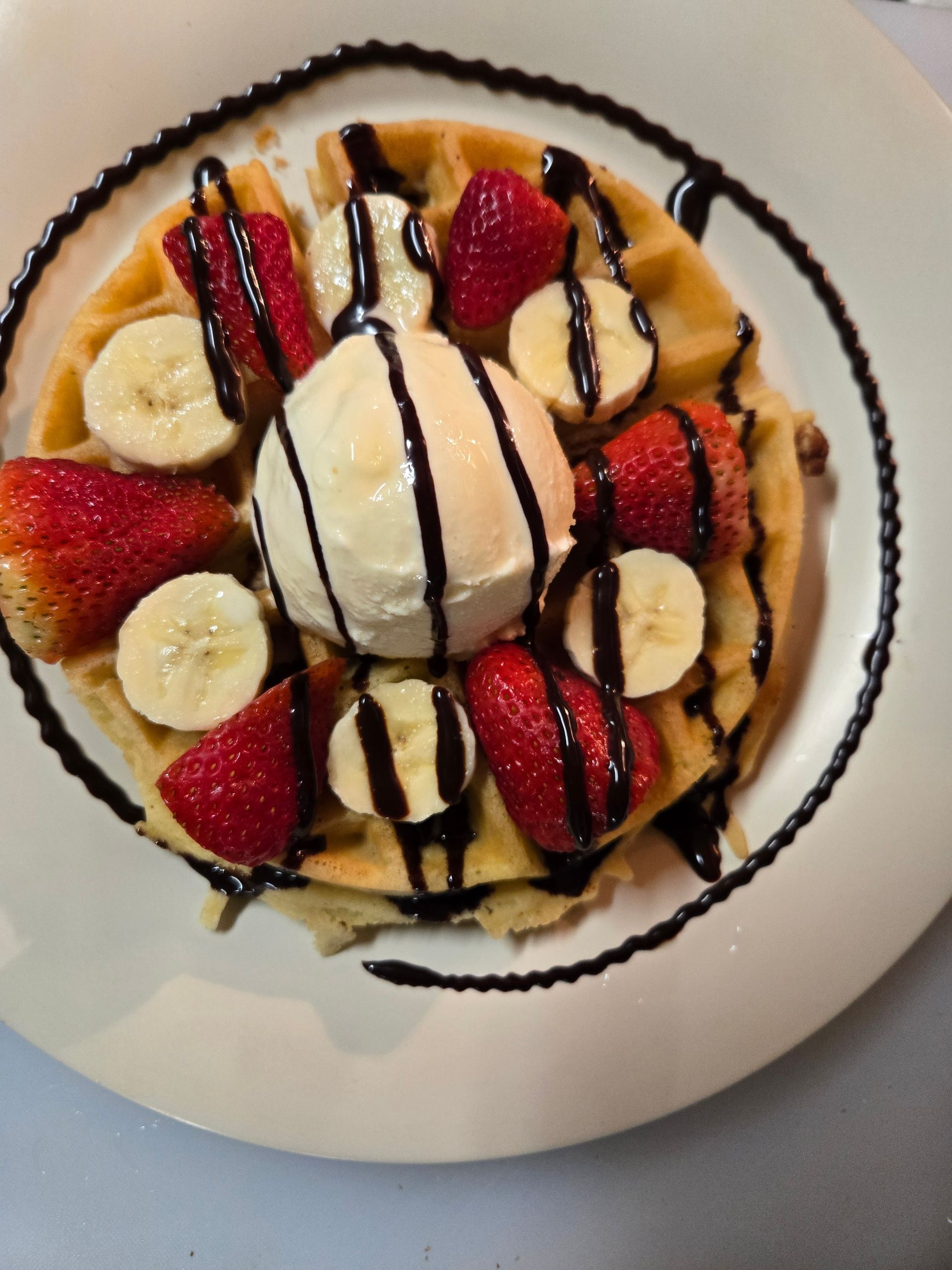 A waffle topped with a scoop of vanilla ice cream, fresh strawberry slices, banana rounds, and a chocolate drizzle.