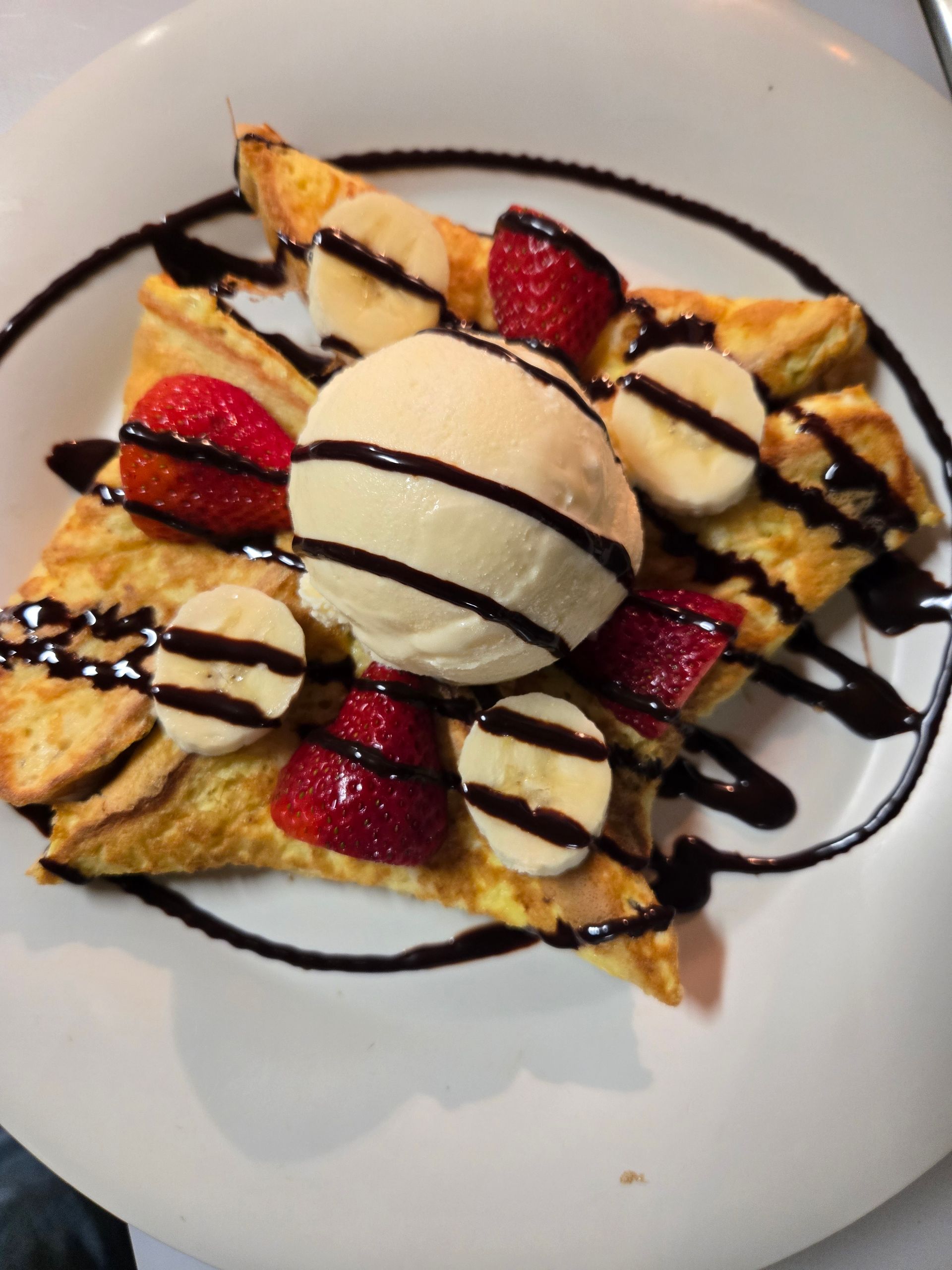 French toast topped with vanilla ice cream, strawberry slices, banana rounds, and a chocolate sauce drizzle.