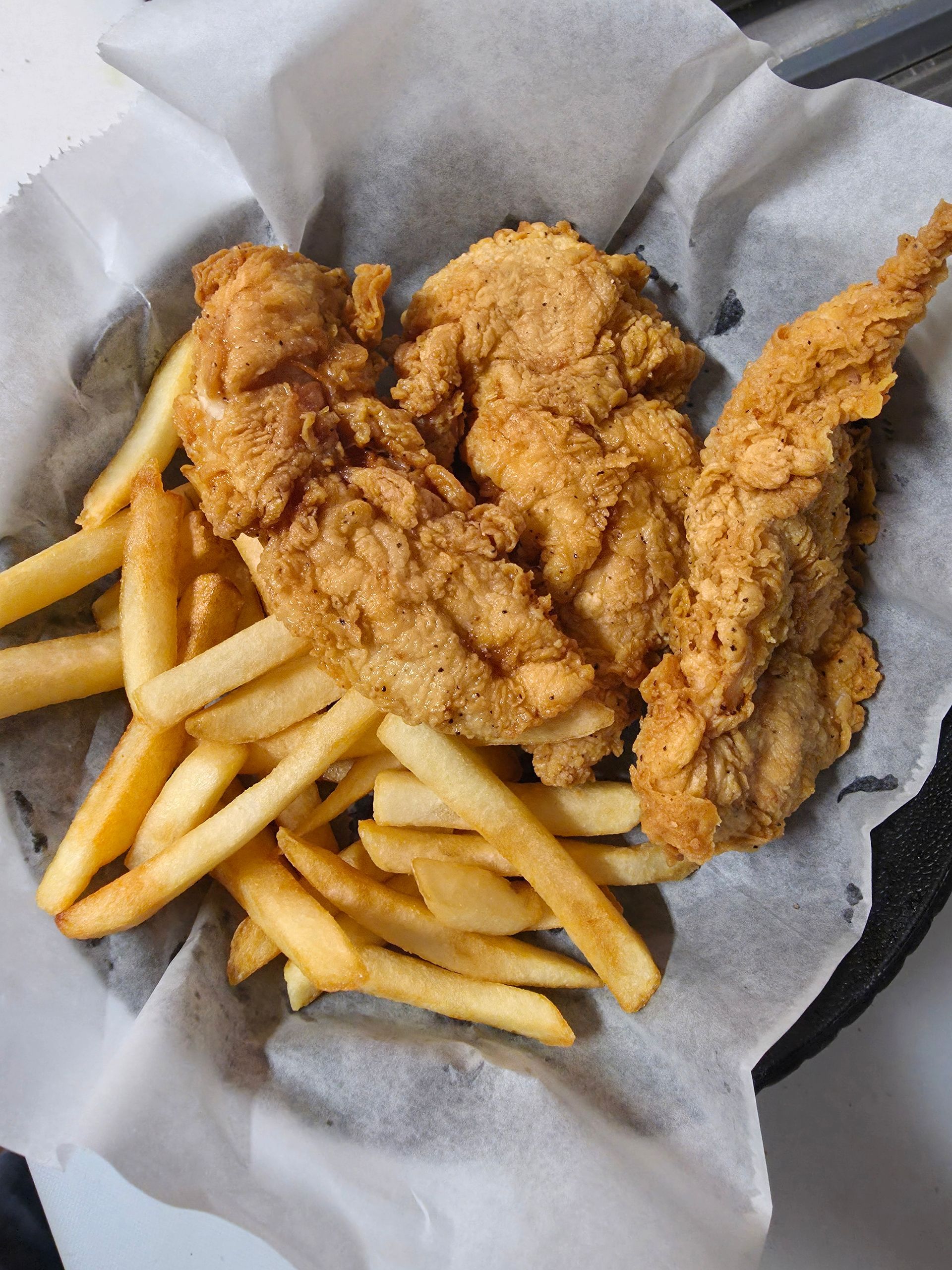 A basket of crispy, golden-brown fried chicken tenders served with a side of shoestring french fries on parchment paper.
