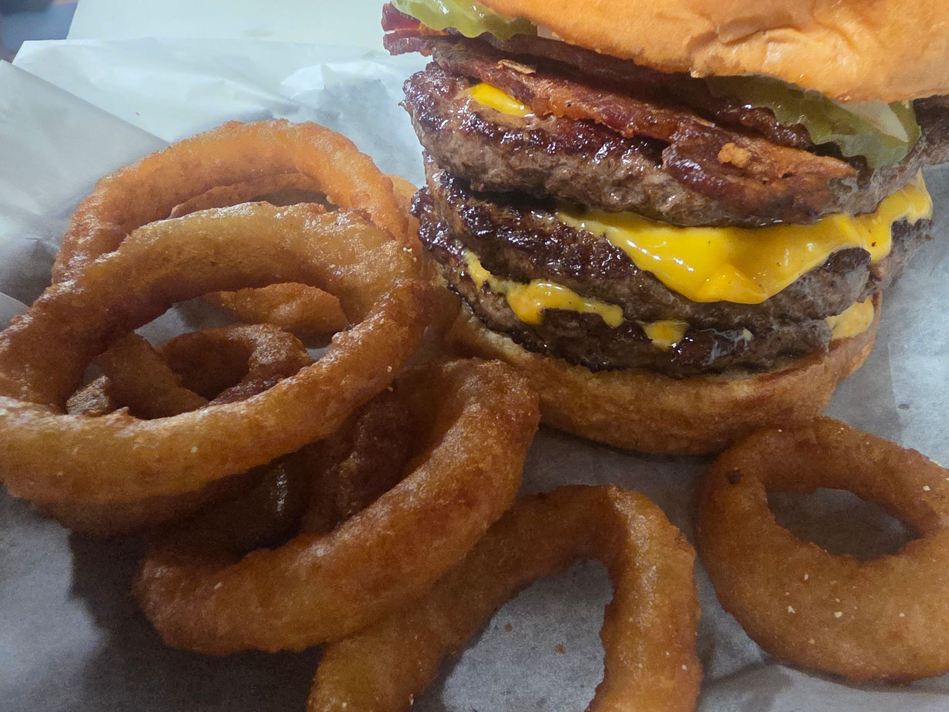A double cheeseburger with bacon and pickles served on parchment paper alongside several golden fried onion rings.