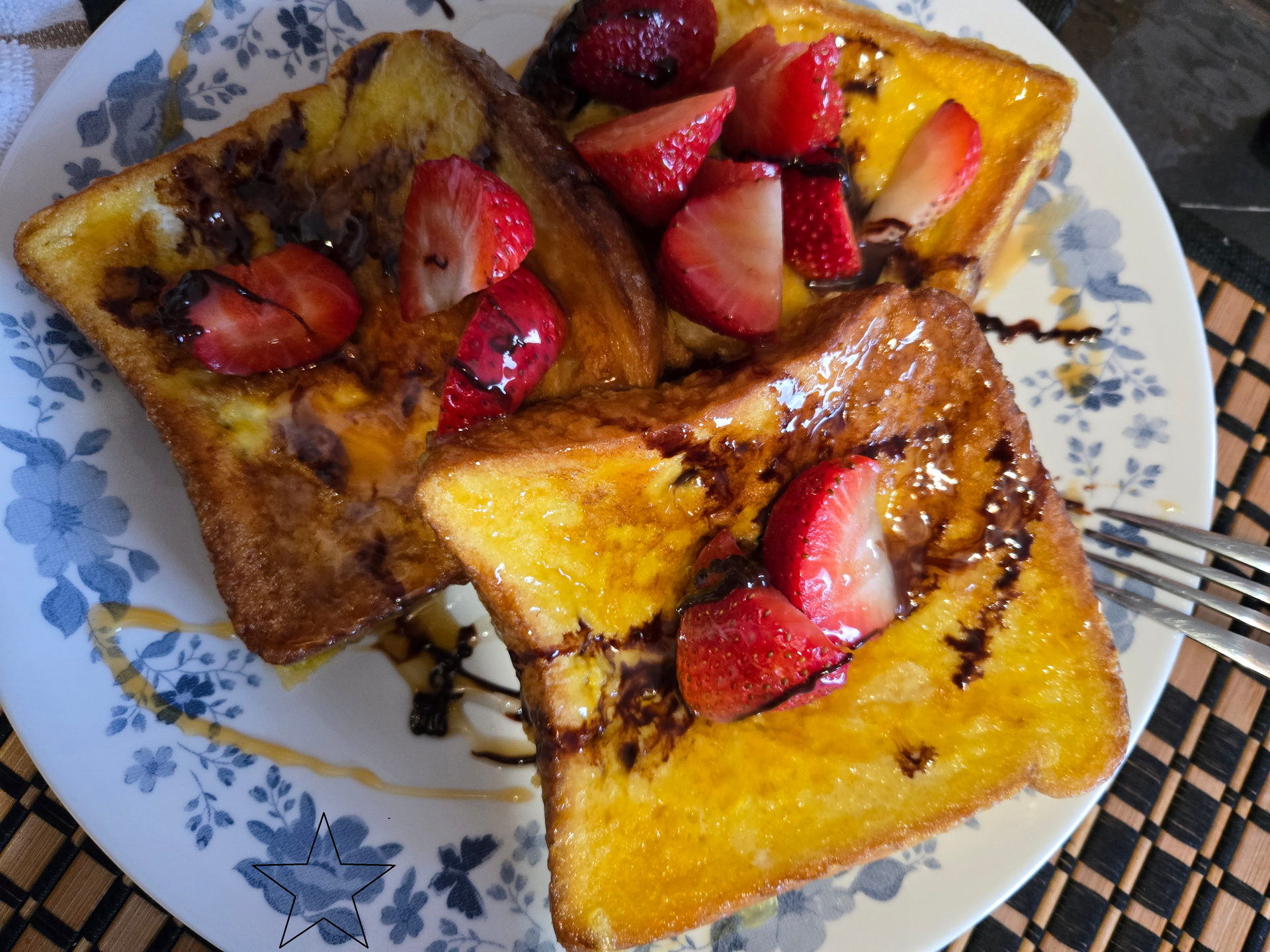 Two pieces of golden-brown French toast on a floral plate, topped with fresh sliced strawberries and chocolate drizzle.