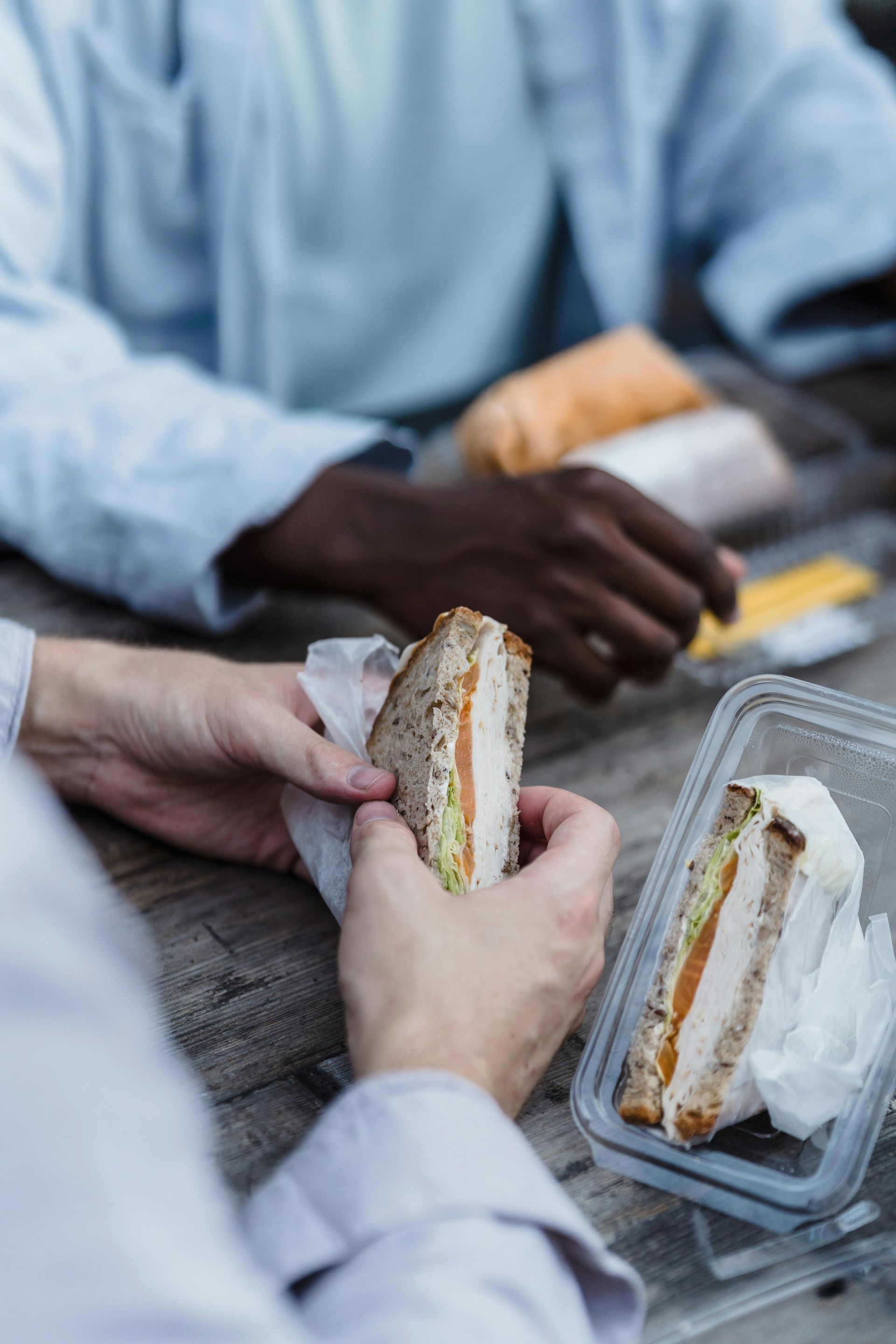 Two individuals sitting at a wooden table holding and preparing to eat sandwich halves from clear plastic containers.