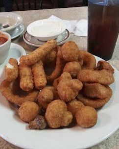 A plate of fried appetizers including mozzarella sticks, onion rings, and breaded mushrooms with dipping sauces and a drink.