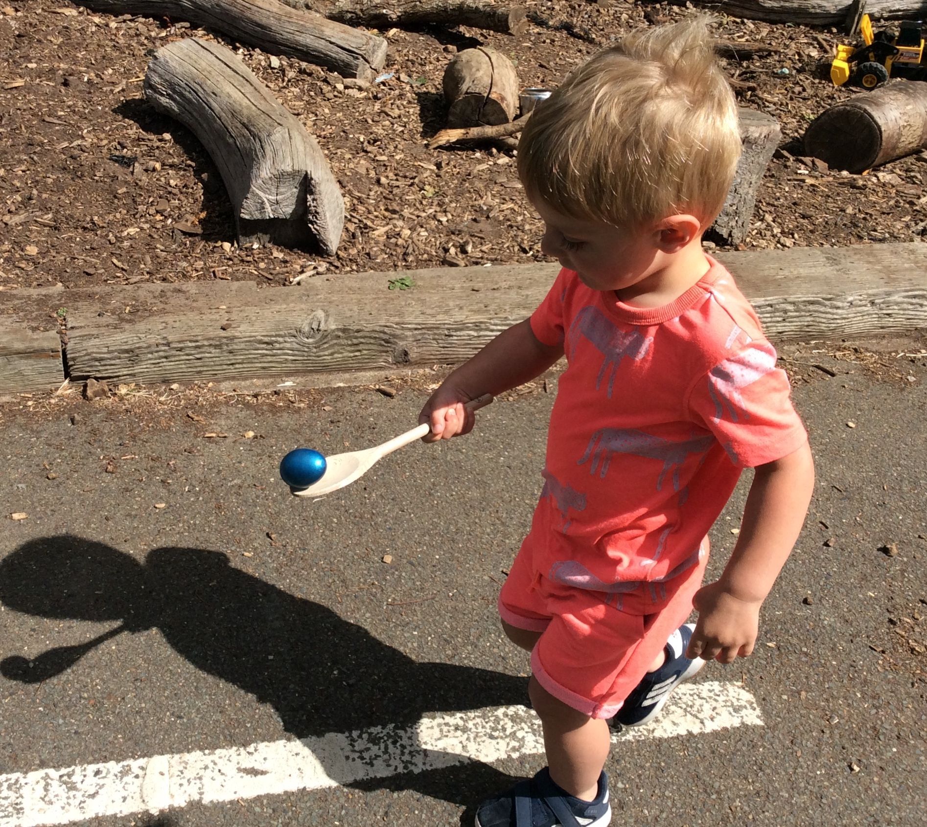 A young boy in a pink shirt is holding a blue ball and a stick