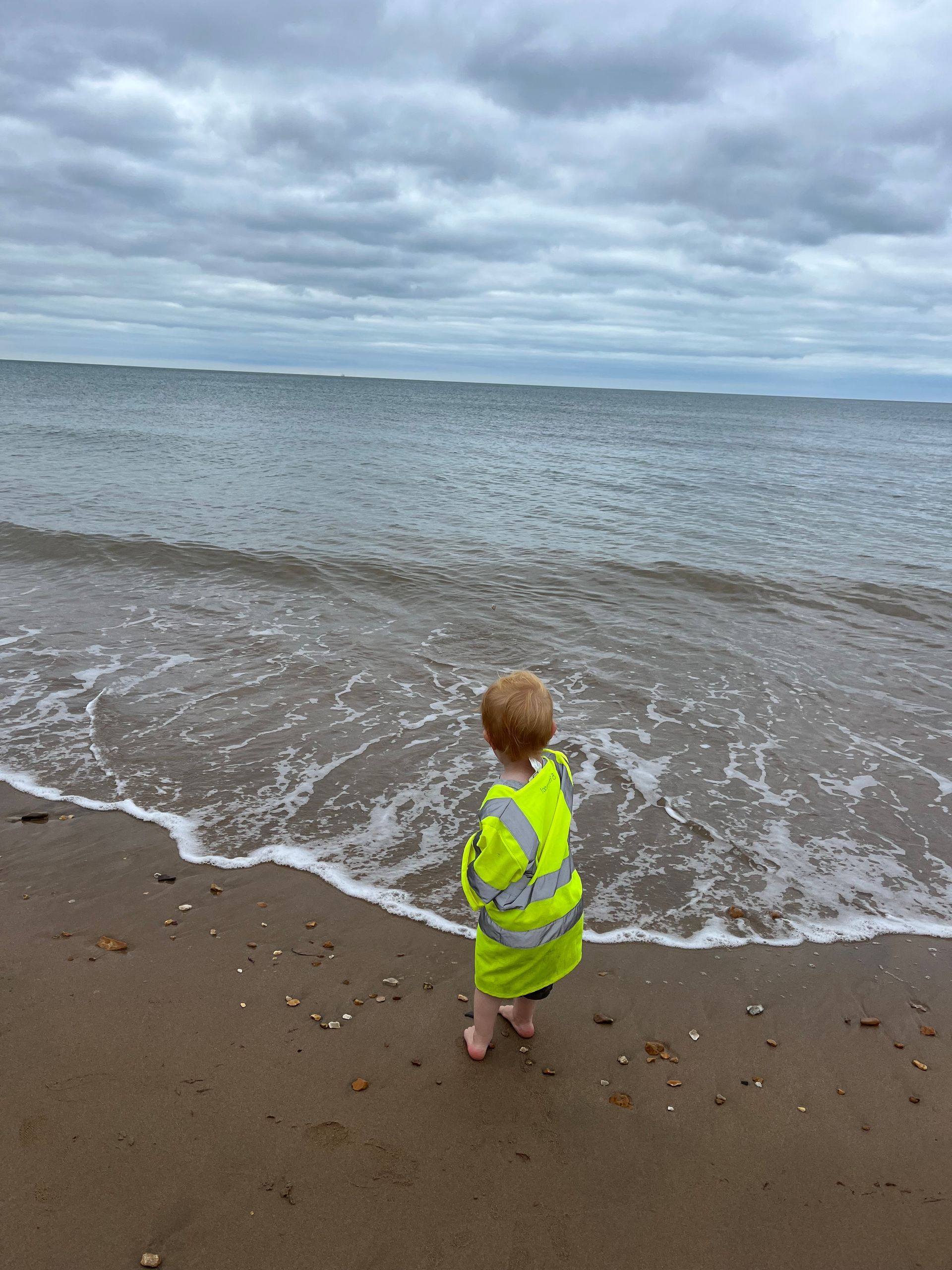 A little boy in a yellow jacket is standing on a beach looking at the ocean.