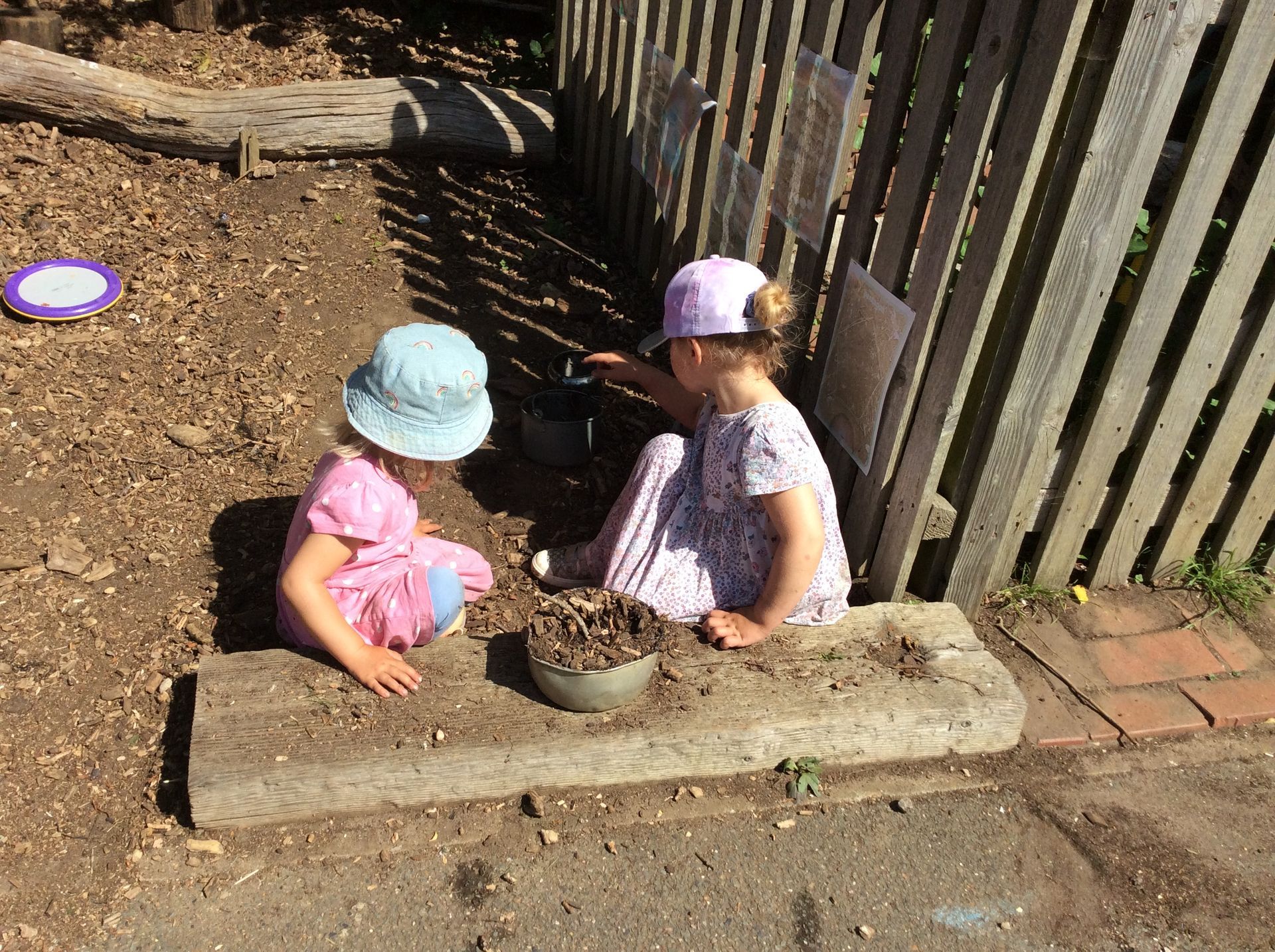 Two little girls are playing in the dirt near a fence.