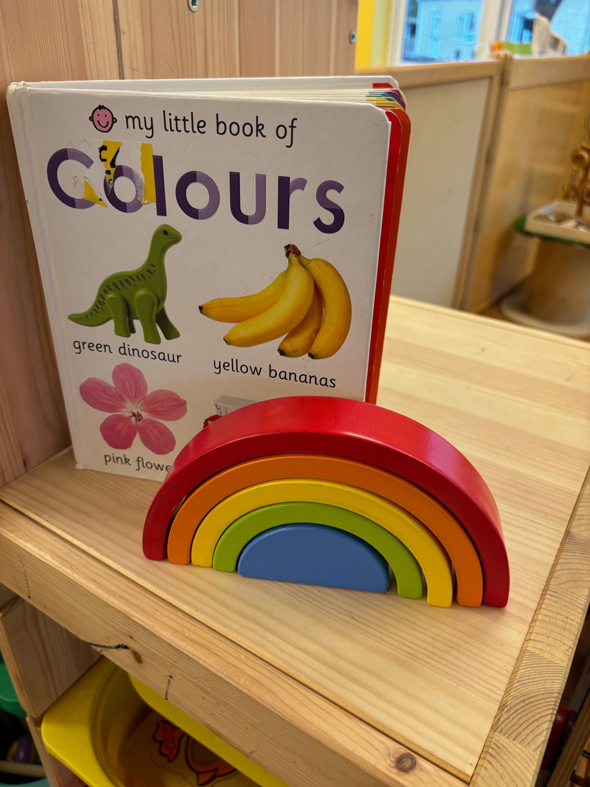 A little book of colors is sitting on a wooden shelf next to a wooden rainbow.