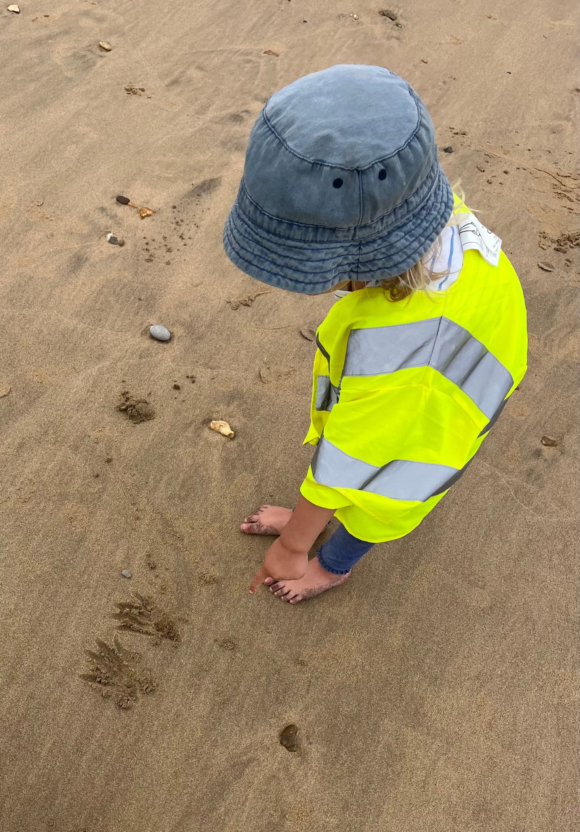 A little boy wearing a hat and a yellow jacket is playing in the sand on the beach.
