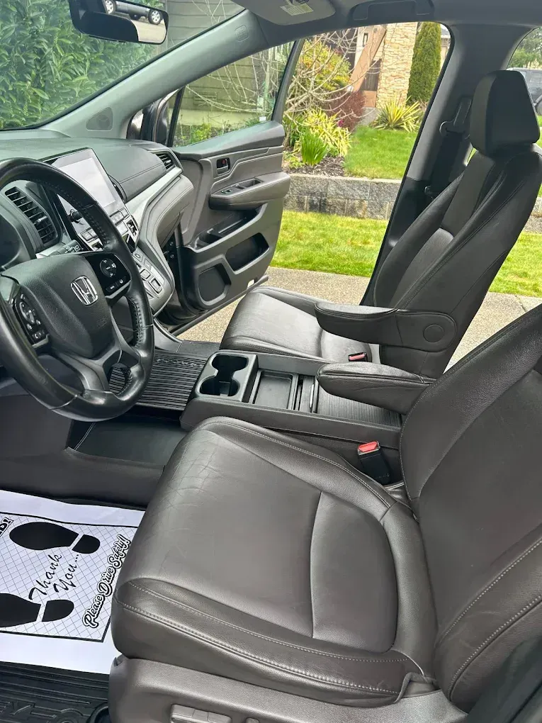 Car interior with black leather seats and steering wheel, front passenger door open to a garden outside