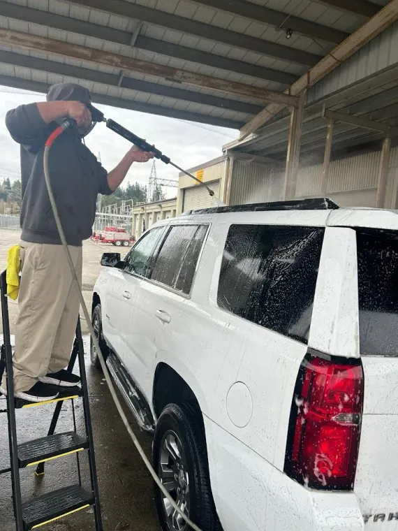 Person pressure-washing a white SUV under a covered car wash area.