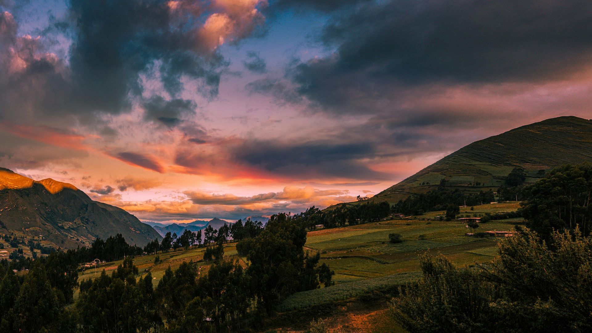 A sunset over a mountain range with trees in the foreground and a cloudy sky in the background.