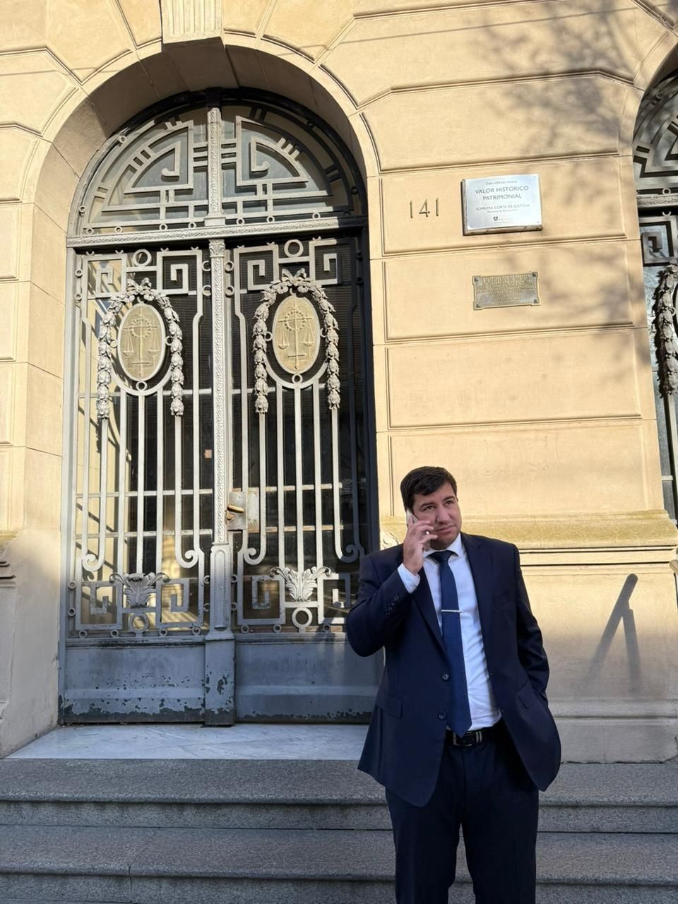 Un hombre con traje oscuro usa un teléfono en los escalones de la entrada de un edificio ornamentado.