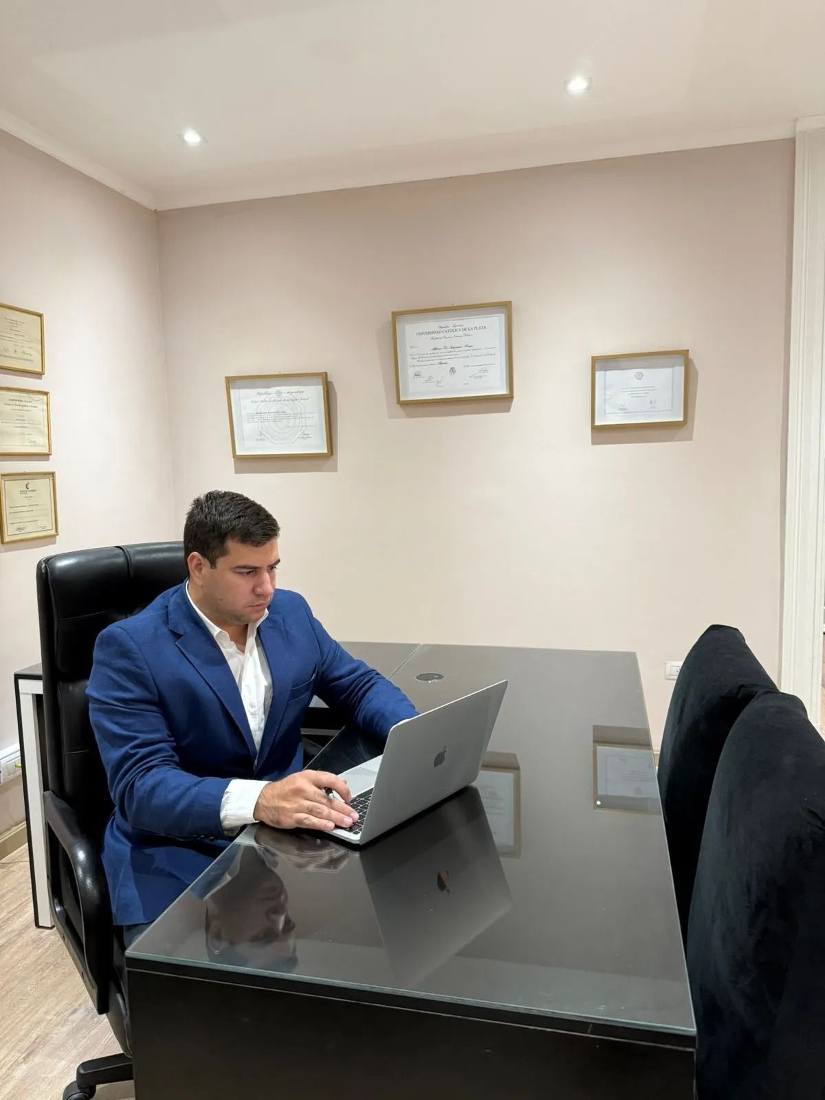 Hombre con traje azul trabajando en un portátil en una mesa de conferencias negra en una pequeña oficina.