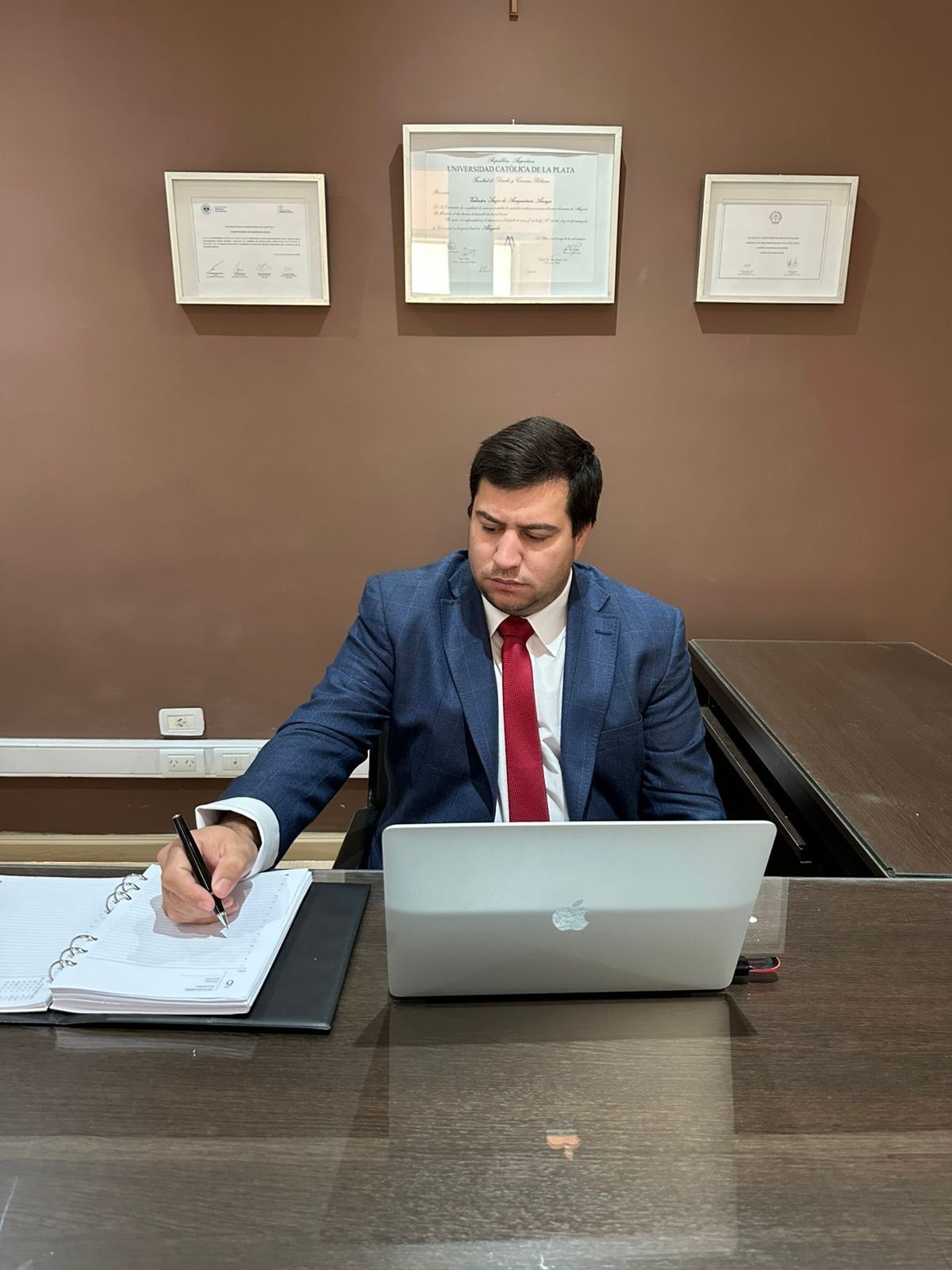 Hombre con traje azul y corbata roja escribiendo en un escritorio con una computadora portátil en una oficina marrón.