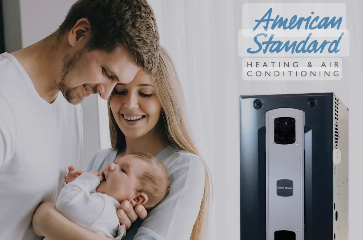 A man and woman are holding a baby in front of an american standard air conditioner.