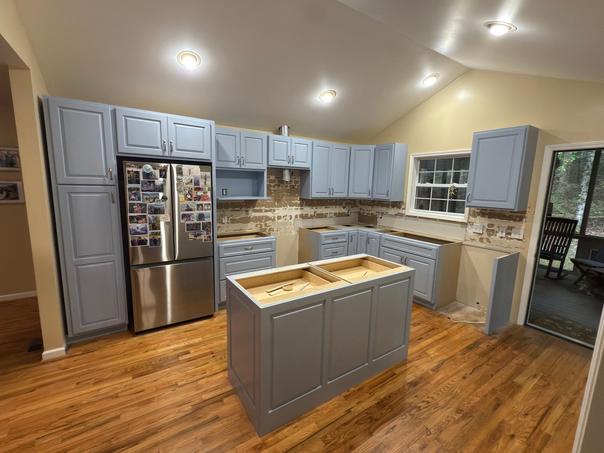 Kitchen renovation: Light blue cabinets, stainless steel refrigerator, island, exposed walls, and hardwood floors.