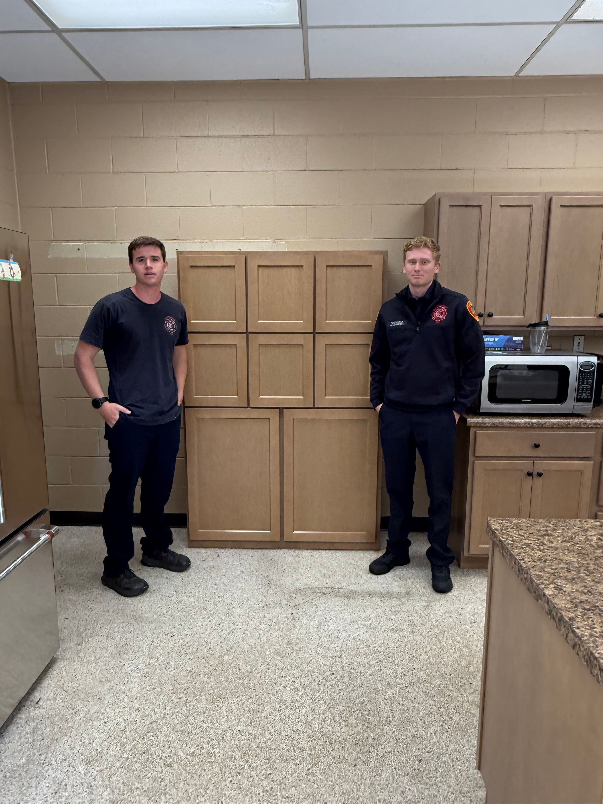 Two firefighters standing in a kitchen with new cabinets. Beige cabinets and wall.