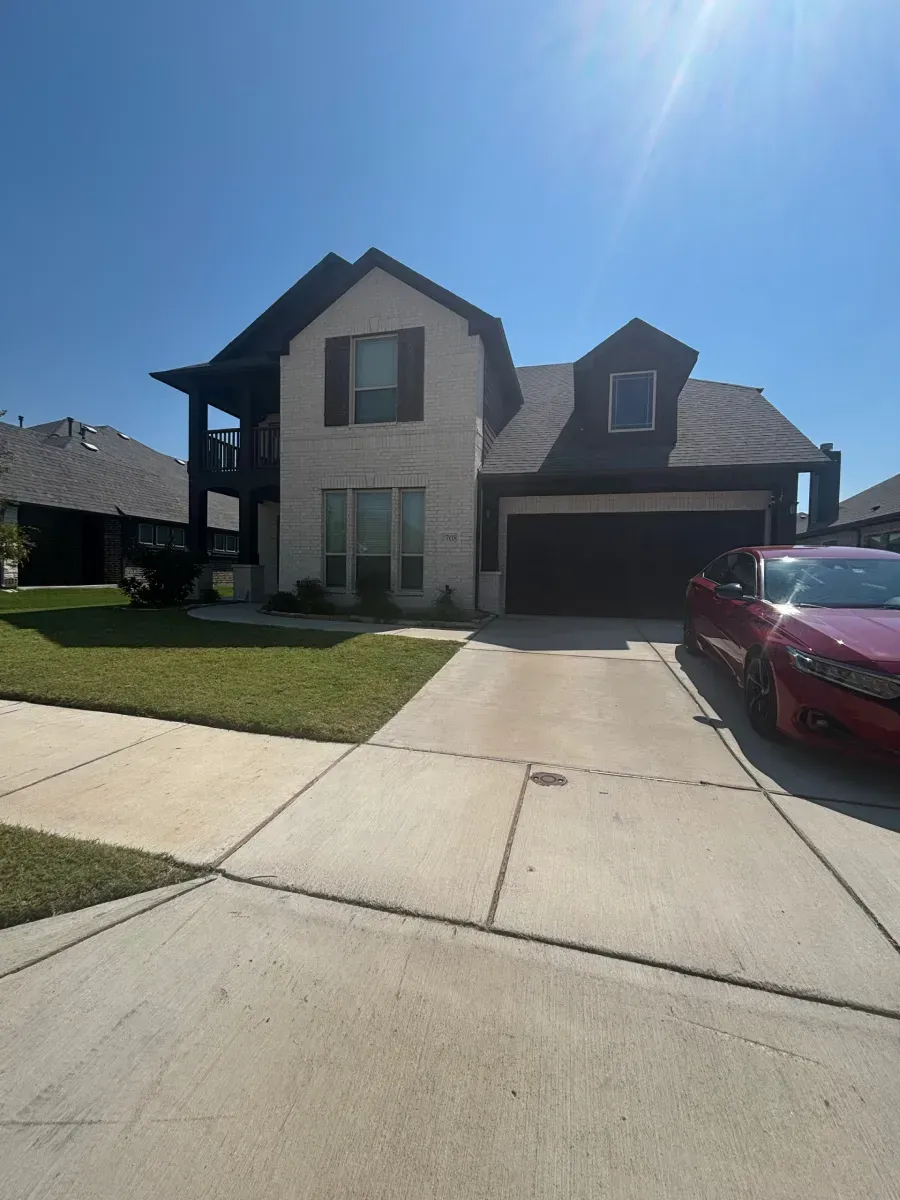 A two-story house with white brick, dark shutters, and a driveway featuring a parked red car under a bright, sunny sky.
