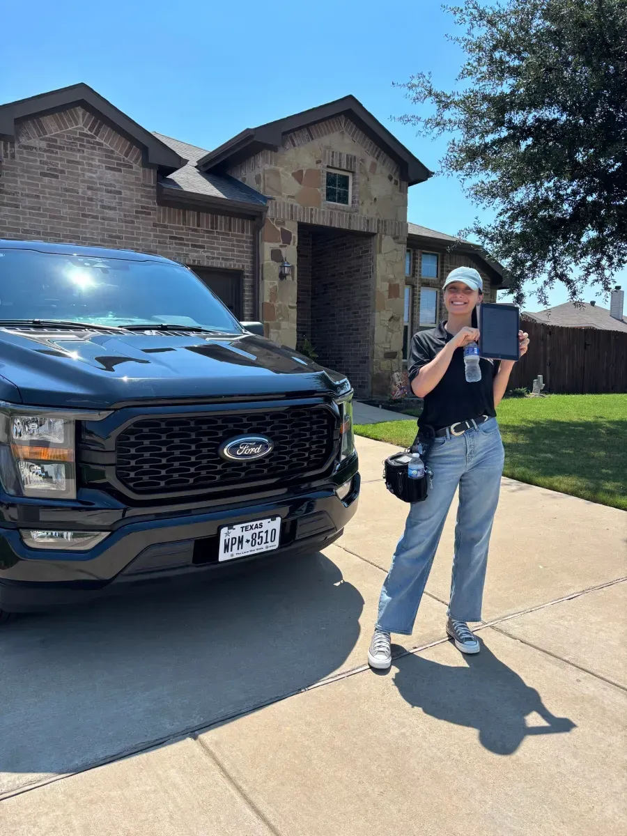 A person stands in a driveway next to a black Ford pickup truck, smiling and holding a tablet in front of a brick house.