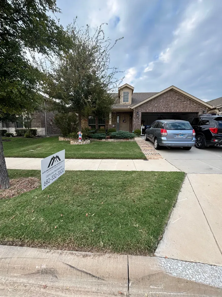 A brick single-story suburban home with a two-car garage, a front lawn with a sign, and two parked SUVs.