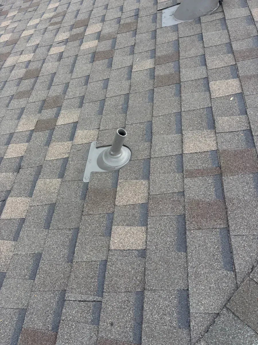 A metal plumbing vent pipe with a flashing base protruding through gray textured asphalt shingles on a sloped roof.