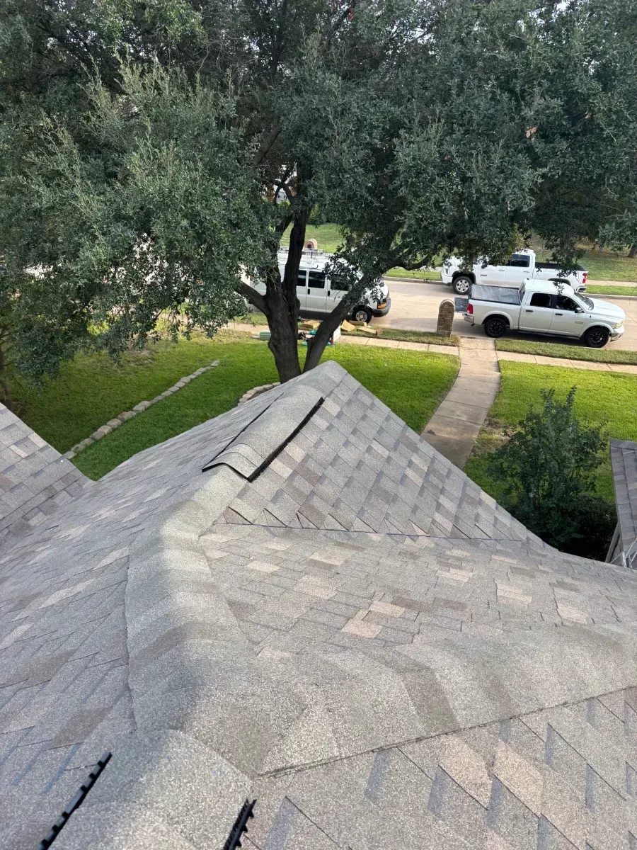 A high-angle view of a gray shingled roof with a vent, overlooking a yard with a large tree and two white pickup trucks.