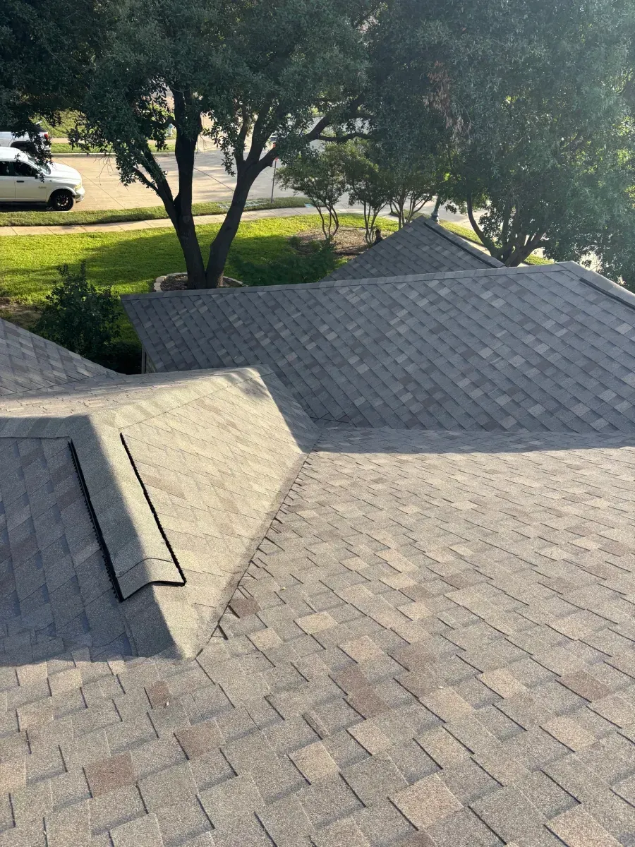 High-angle view of a grey shingled roof with a skylight, surrounded by green trees and a white vehicle in the distance.