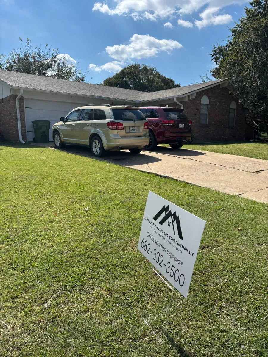 A single-story suburban brick house with two cars parked in the driveway and a real estate sign in the foreground lawn.
