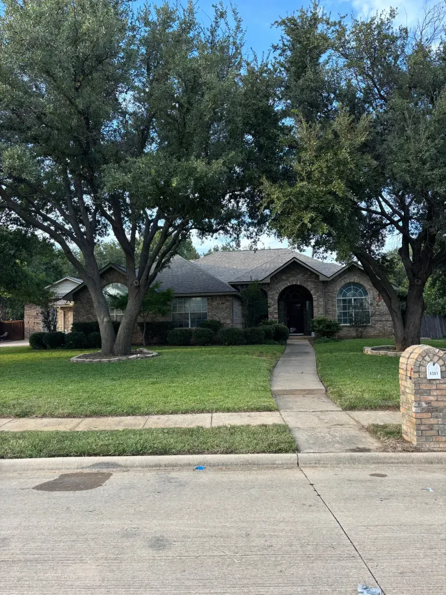 A single-story brick house with a arched entryway, surrounded by mature trees, a green lawn, and a paved walkway.