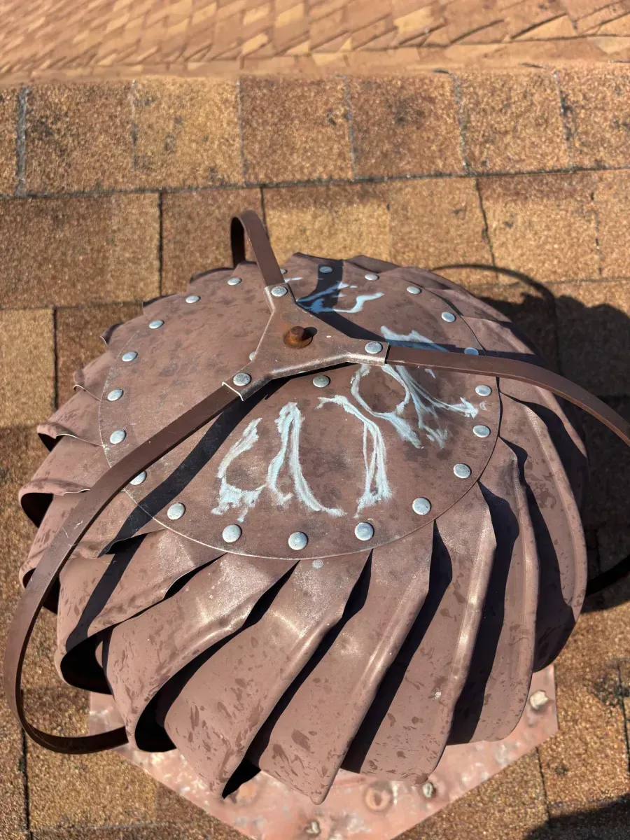 A metal wind turbine roof vent is installed on a brown shingled roof.