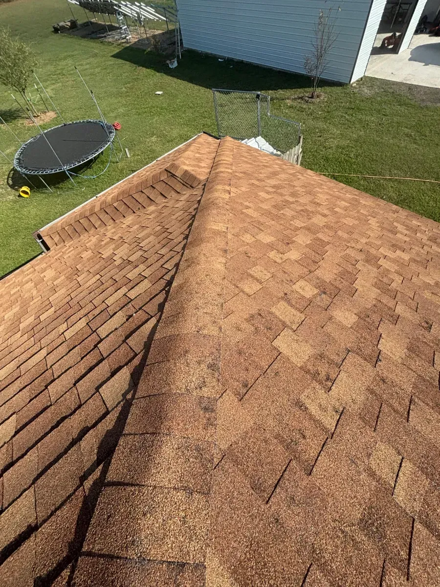 A high-angle view of a brown asphalt shingle roof on a house, with a backyard and trampoline visible in the distance.