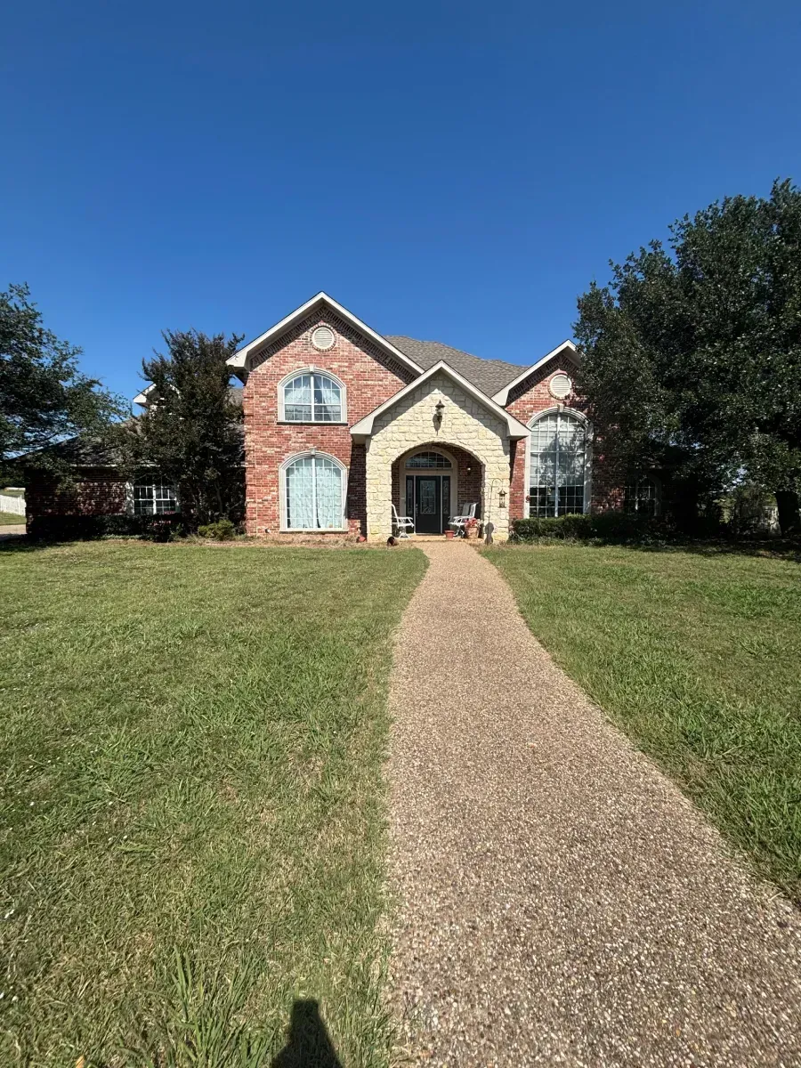 A two-story red brick house with a stone-accented arched entryway, a gravel walkway, and large green trees under blue sky.