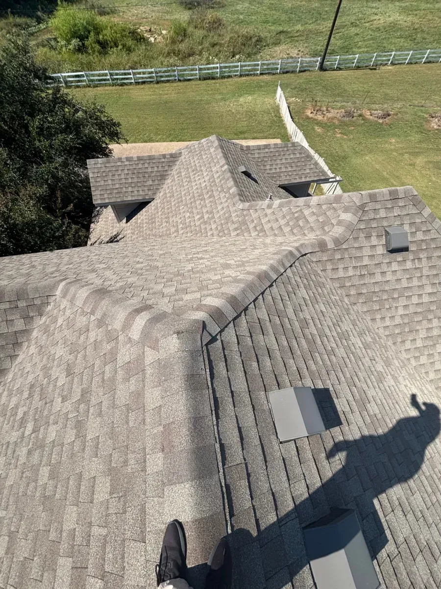 A high-angle view of a gray asphalt shingle roof with visible vents and a person's feet standing at the bottom.