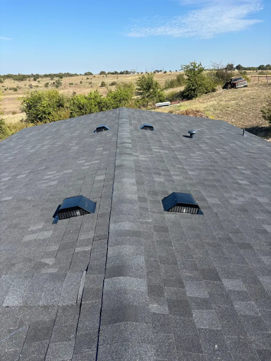 A gray shingled roof with several black attic vents, viewed from above looking down toward a rural landscape.