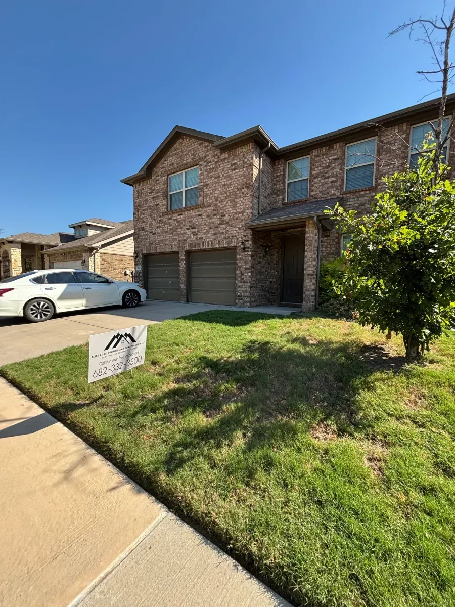 A two-story brick house with a two-car garage, a white car in the driveway, and a small yard sign on a sunny day.