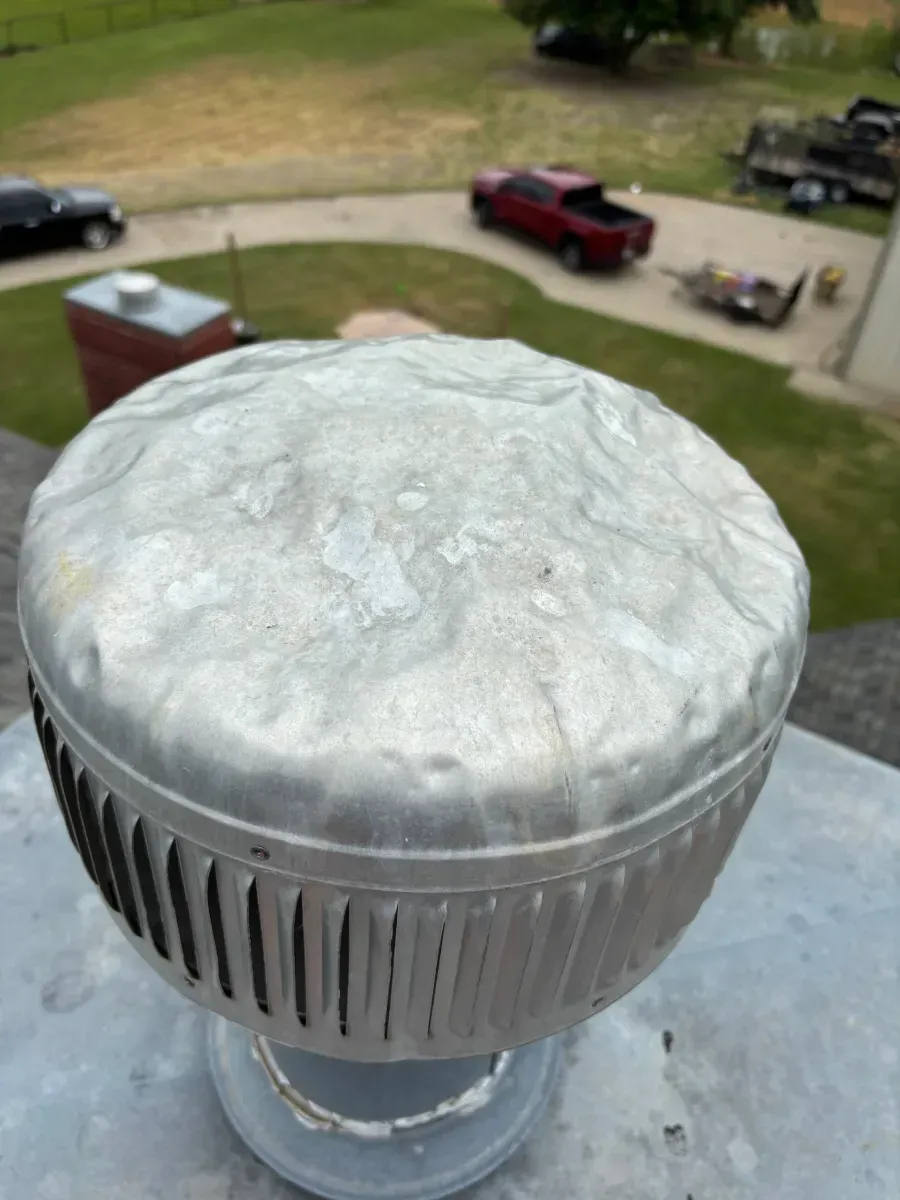 A dented, silver metal vent cap sits on a roof with a driveway and parked vehicles in the blurred background.