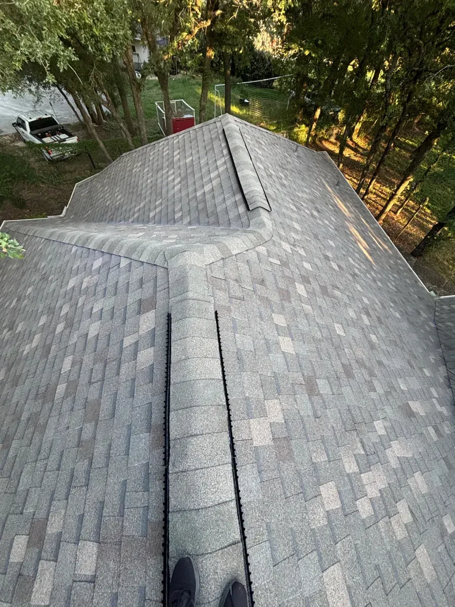 A high-angle view of a gray asphalt shingled roof with a central ridge, surrounded by trees and a glimpse of a vehicle.
